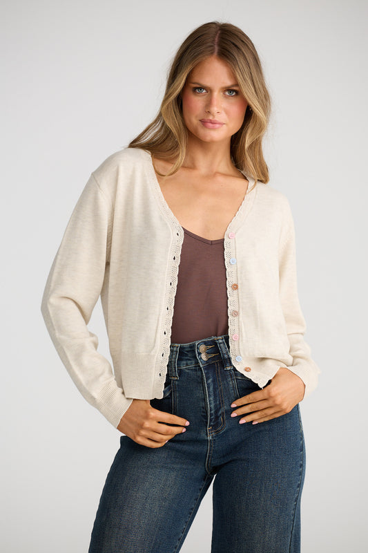 Woman wearing a cream cardigan over a brown top and blue jeans on a white background