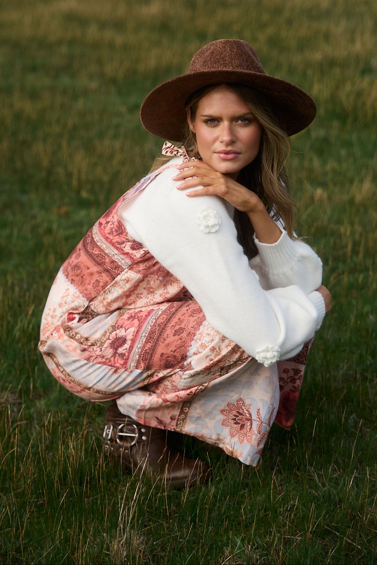 Woman in a patterned dress and wide-brimmed hat sitting in a grassy field.