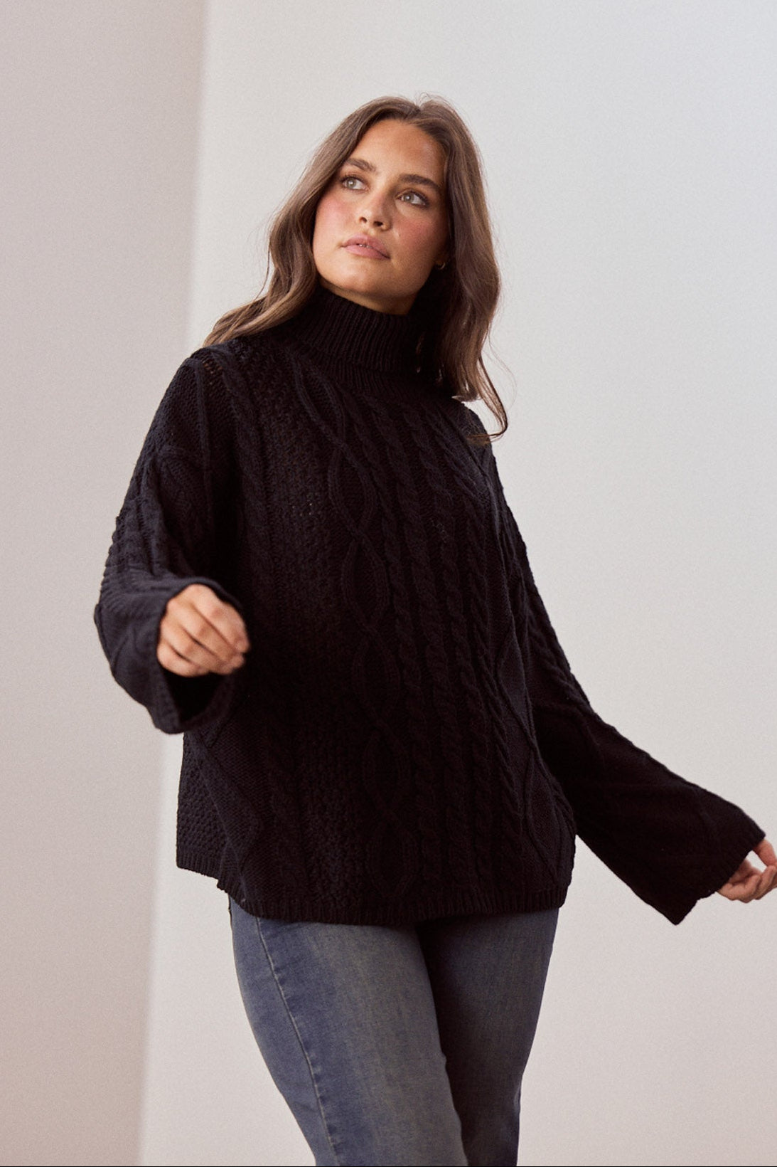 Model wearing Dixie Knit in black with roll-neck collar and cable knit design, side view on white background