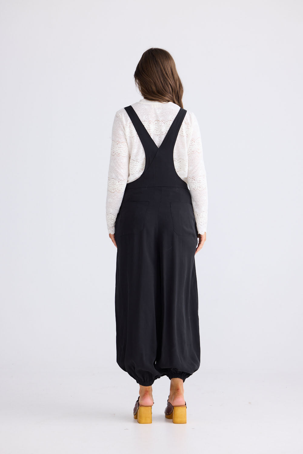 Model wearing Genie Overalls in black with adjustable straps with antique brass button closure, side view on white background
