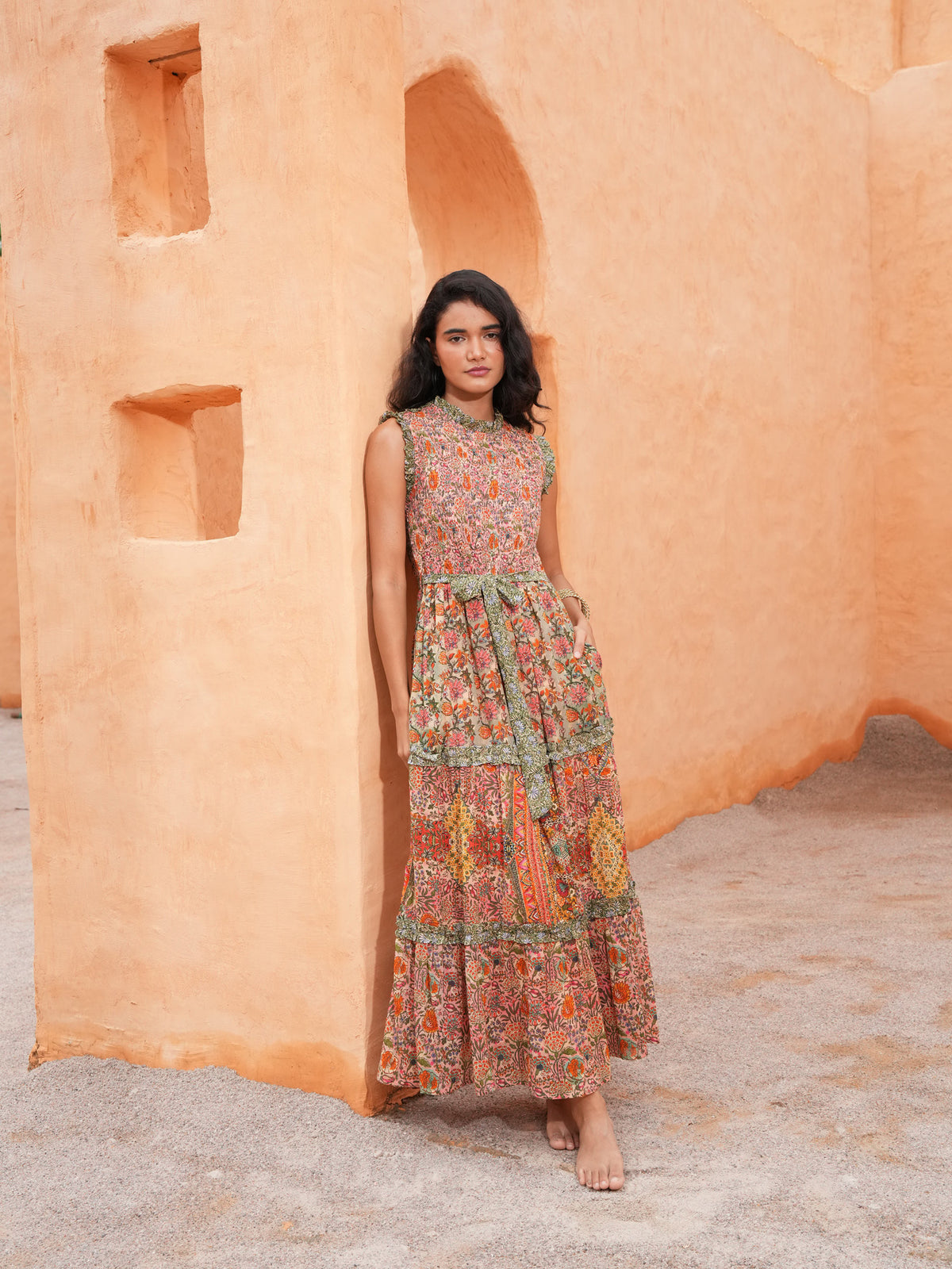 A woman stands barefoot against a warm terracotta stucco wall, wearing a sleeveless, tiered maxi dress with an intricate multicolor floral and paisley print in orange, green, and beige tones.
