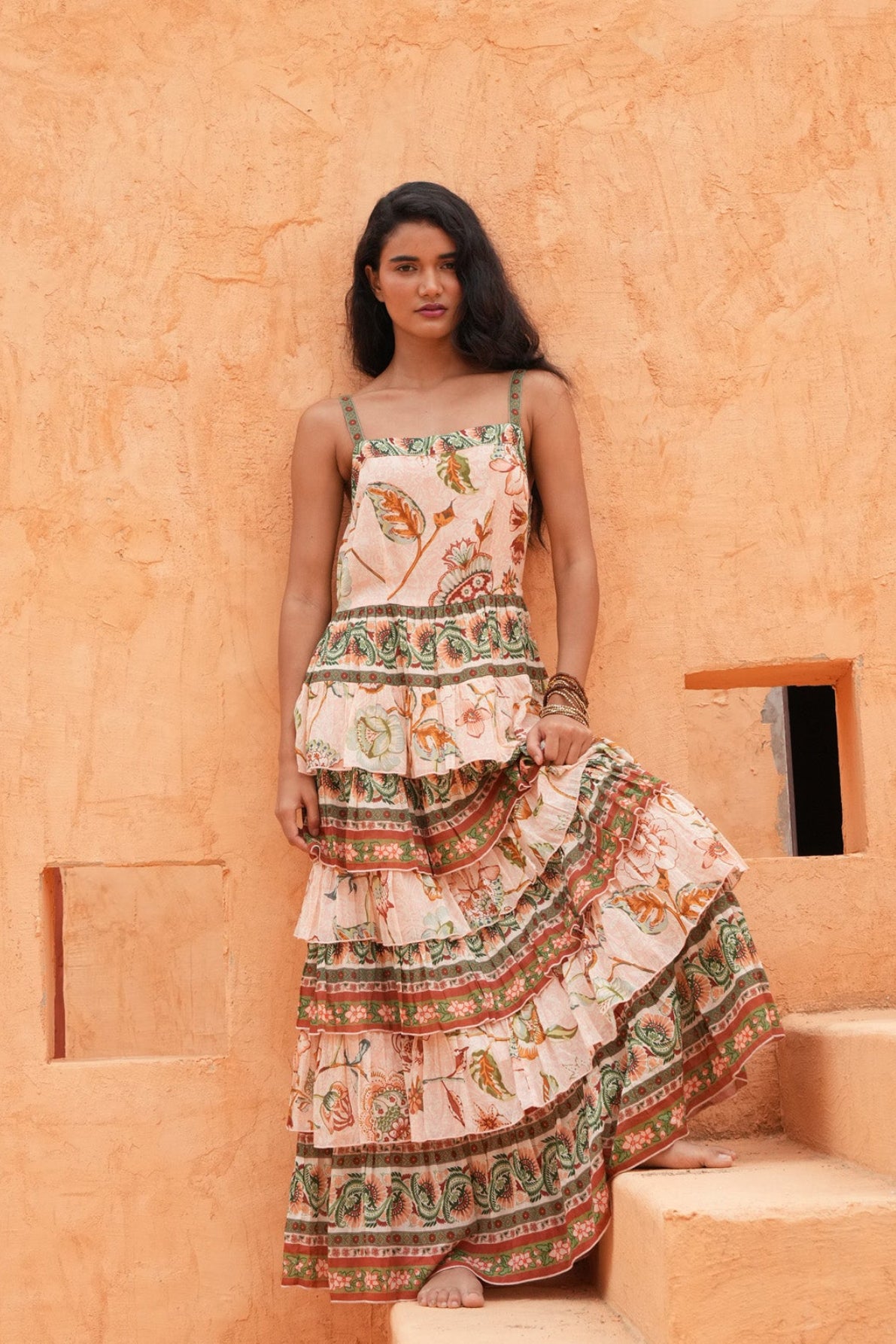 A woman stands on outdoor stone steps in front of a textured orange adobe wall, wearing a sleeveless tiered maxi dress with a cream base and colorful botanical and geometric patterns.