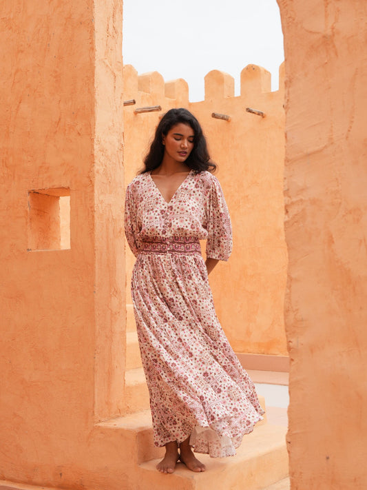 A woman wearing a long, loose floral maxi dress stands barefoot between warm orange stucco walls in an outdoor architectural setting.
