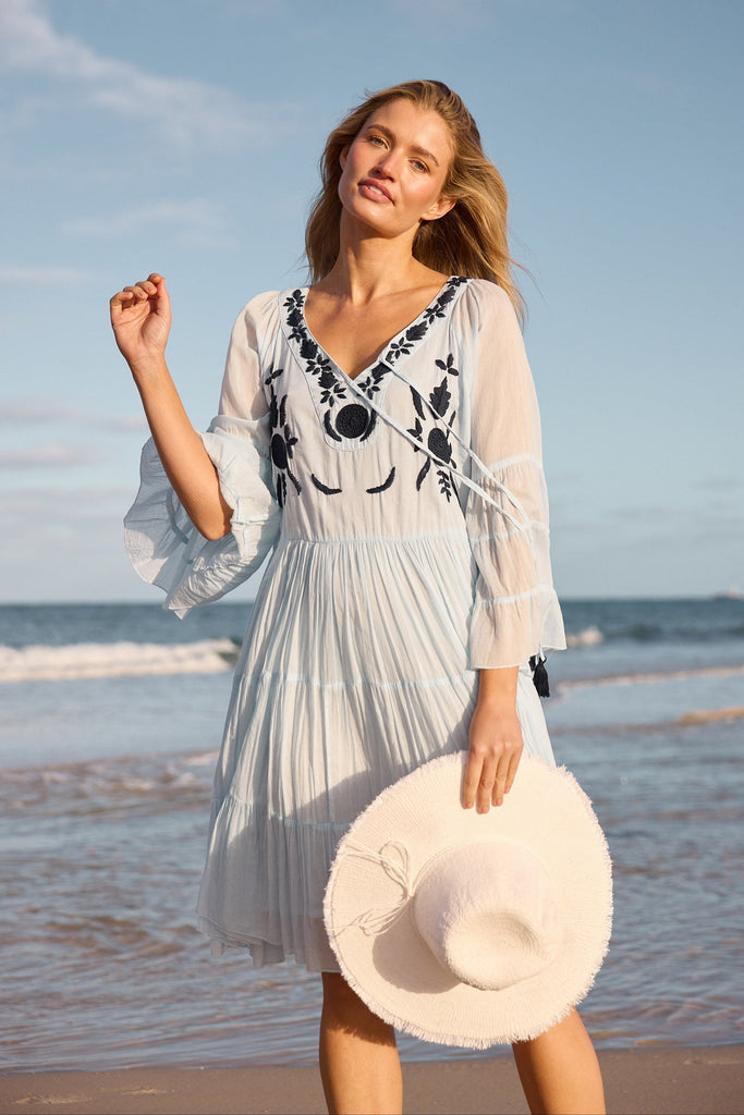 Woman in a light blue dress with navy embroidery on a beach holding white hat