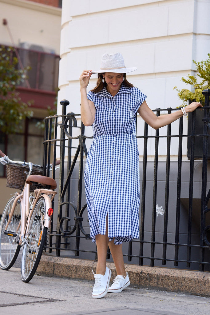 Woman in a blue checkered dress and grey hat standing on a sidewalk with a bicycle in the background.