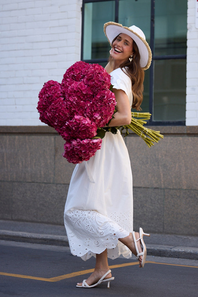Woman in a white dress and hat holding pink flowers against a building background