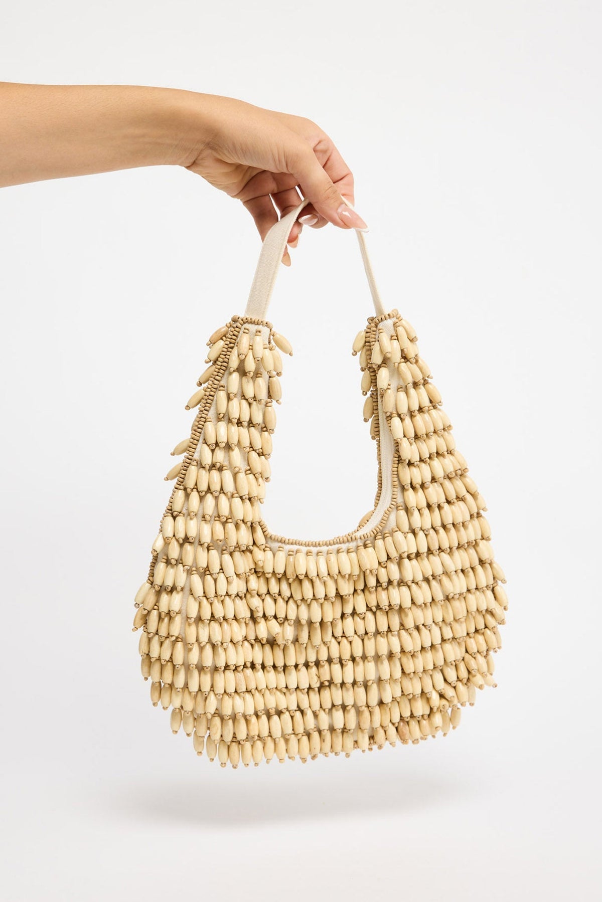 Crescent-shaped shoulder bag fully covered in rows of natural cream oval wooden beads with gold-tone accent beads, held by hand against a white background
