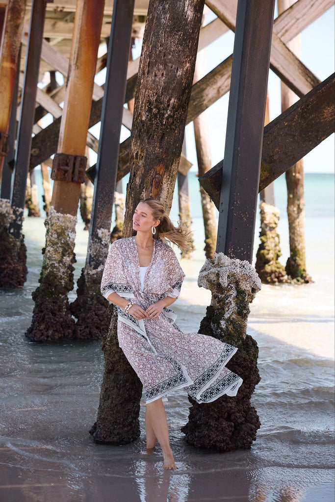 Woman in a patterned kaftan sitting under a wooden pier by the water.