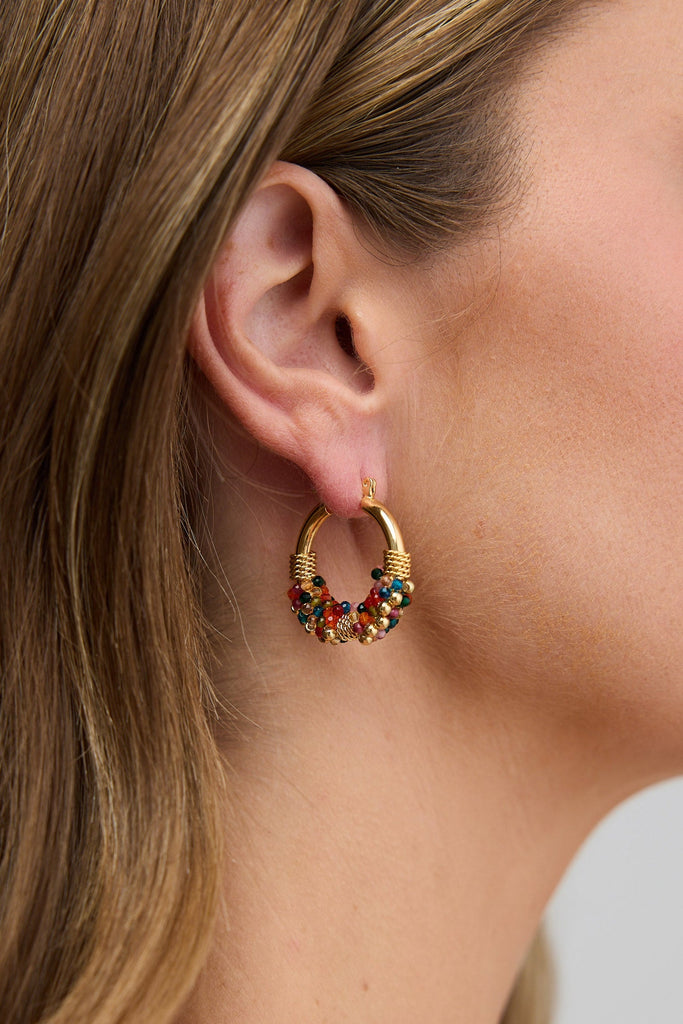 Close-up of a woman wearing gold hoop earrings with colorful gemstones against a neutral background