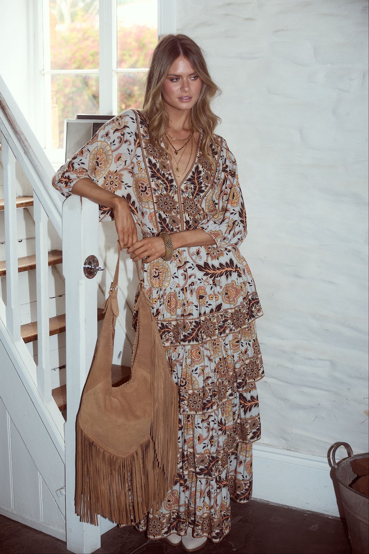 Woman in a floral dress holding a beige handbag with fringe, standing in a home setting.