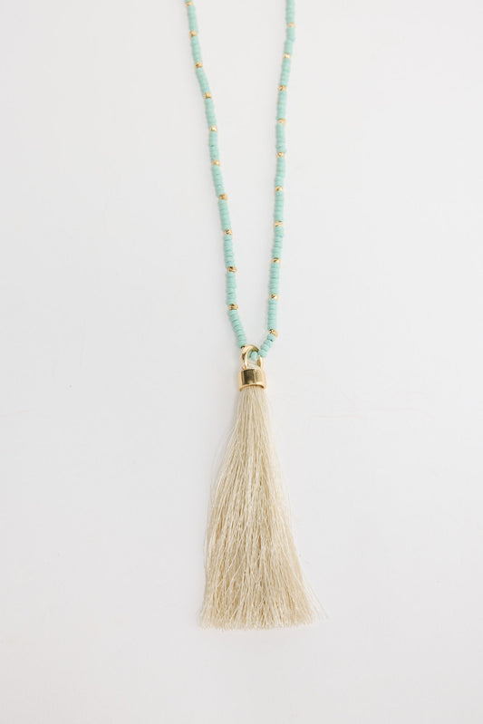 Long aqua-blue seed bead necklace with gold-tone spacer beads and a large cream silk tassel pendant with a gold cap, shown flat on a white background