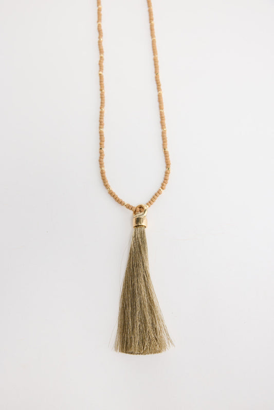 Long neutral taupe seed bead necklace with gold-tone spacer beads and a large olive-gold silk tassel pendant with a gold cap, shown flat on a white background
