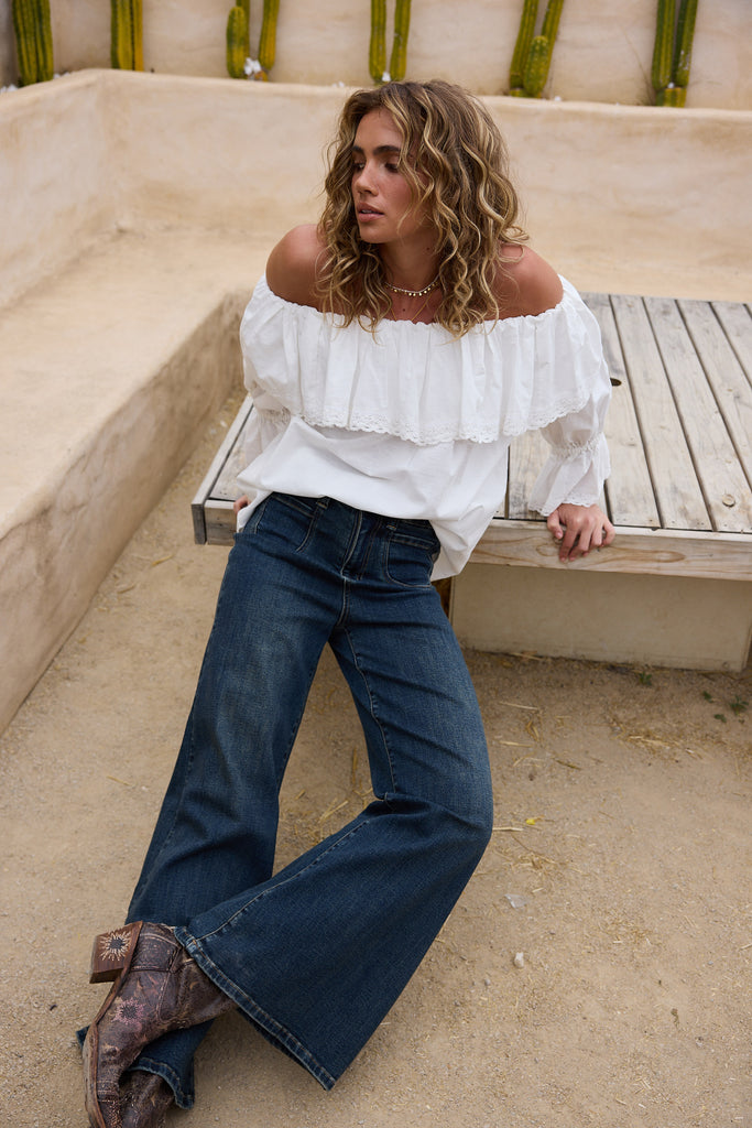 Woman sitting on a wooden bench wearing a white off-shoulder top and blue jeans.