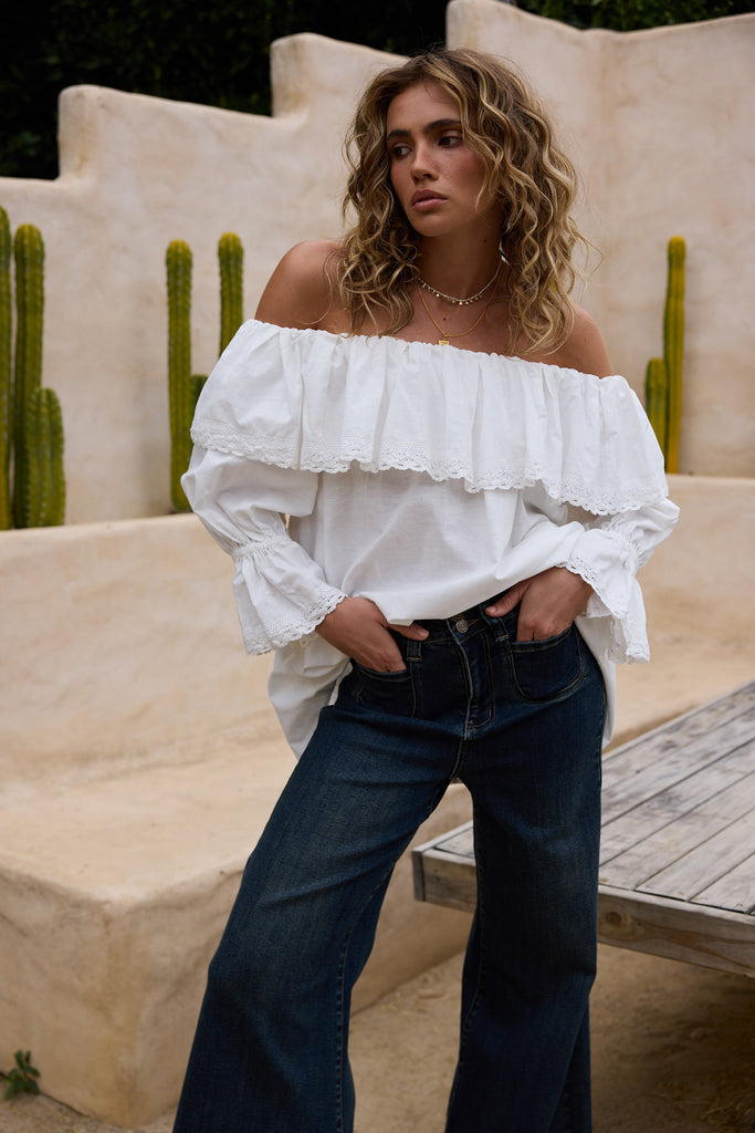 Woman wearing a white off-shoulder top and dark jeans in an outdoor setting with cacti.