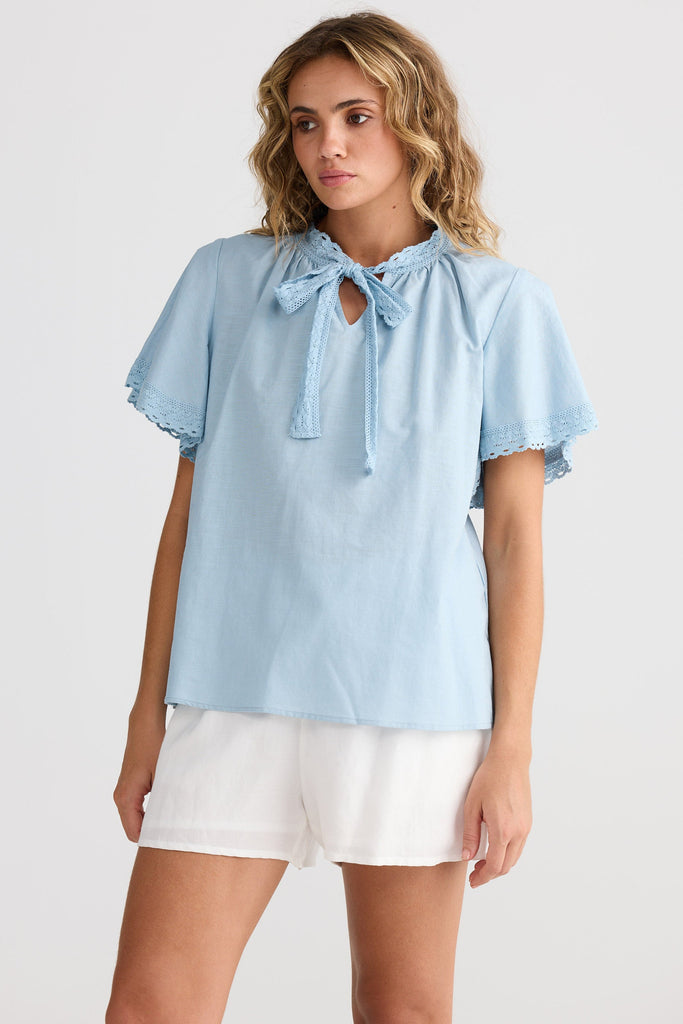 Woman wearing a light blue blouse with ruffled sleeves and white shorts on a plain background