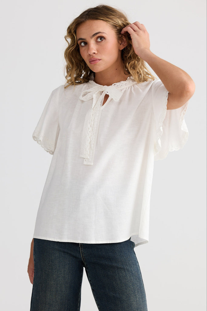 Woman wearing a white blouse with lace details and dark jeans on a plain background