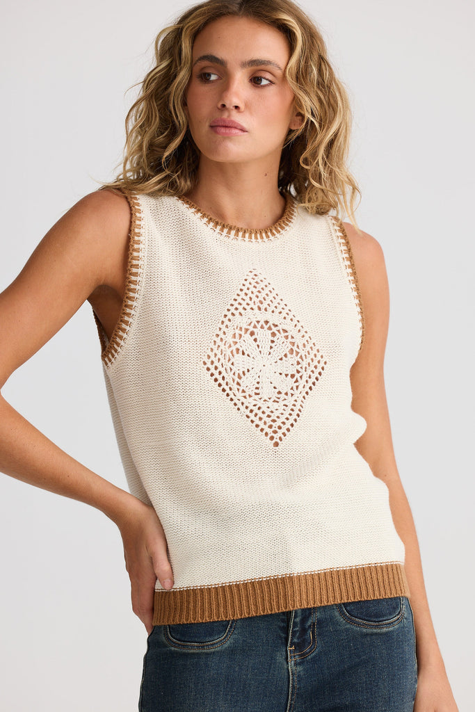 Woman wearing a cream sleeveless top with brown trim and pattern, standing against a plain background.