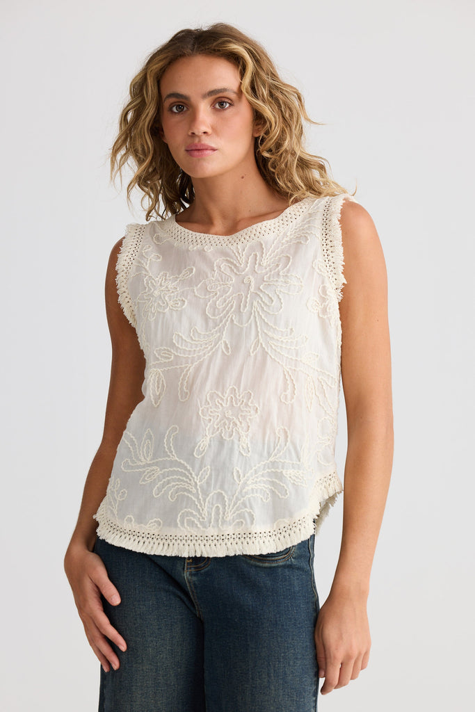 Woman wearing a white sleeveless top with embroidered details against a plain background