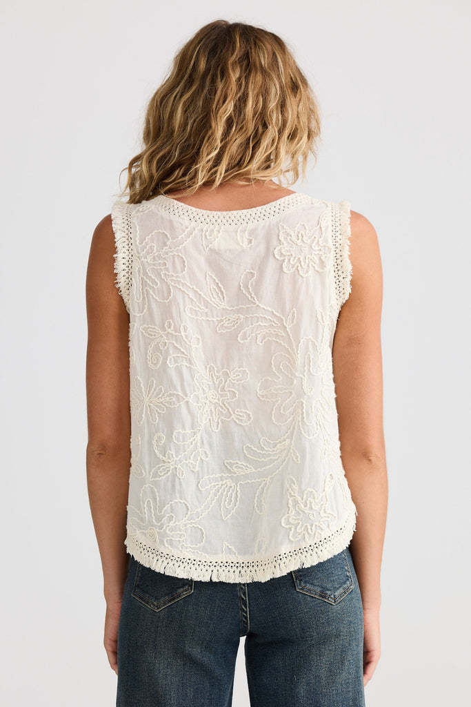 Person wearing a white sleeveless top with embroidered detail and blue jeans on a plain background