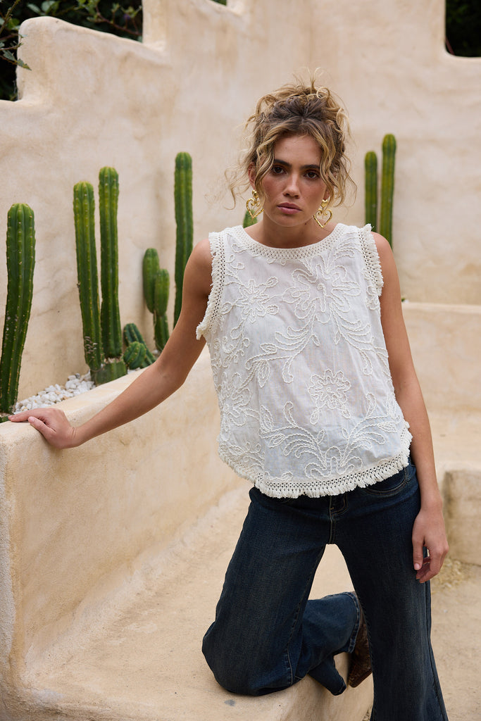 Woman wearing a white top with embroidered detail and dark jeans standing in front of cacti.