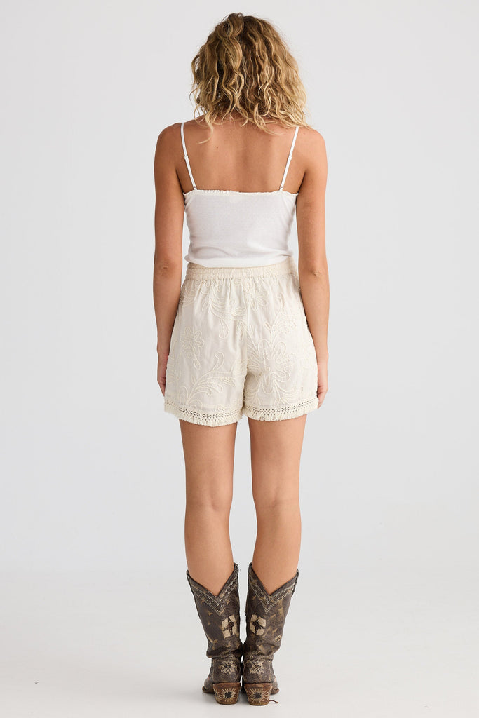 Woman wearing a white tank top and beige shorts with cowboy boots on a white background