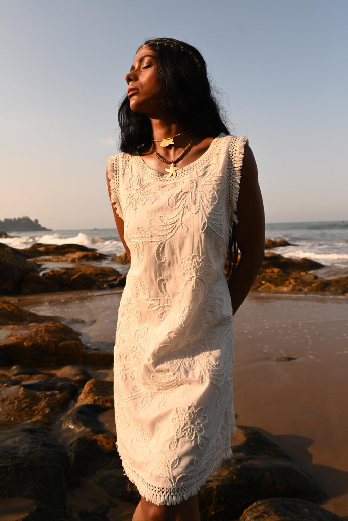 Woman in a white dress standing on a beach with water and rocks in the background