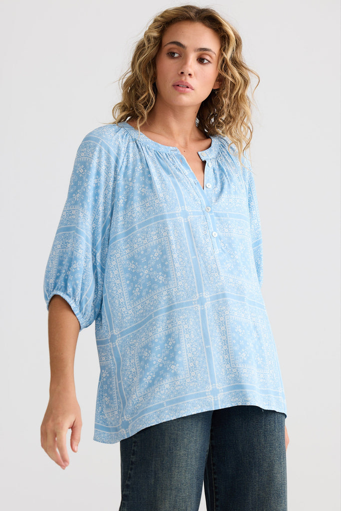Woman wearing a light blue patterned top and dark jeans on a white background