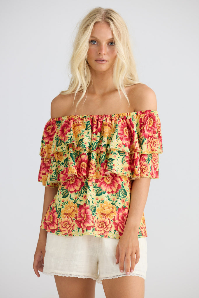 Woman wearing a colorful floral off-shoulder top and white shorts on a plain background