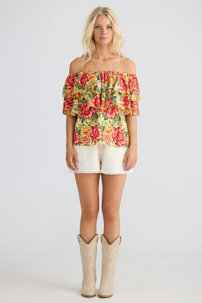 Woman wearing a colorful floral off-shoulder top and white shorts with beige boots on a plain background