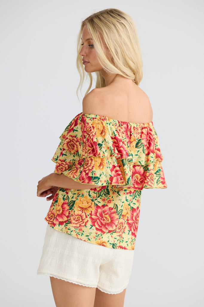 Woman wearing a colorful floral off-shoulder top and white shorts on a plain background