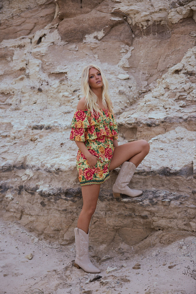Woman in a colorful dress and cowboy boots standing against a rocky cliff.