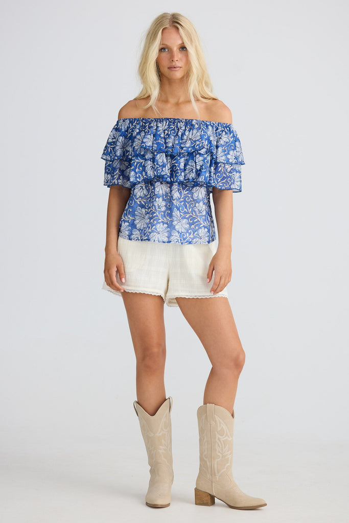 Woman wearing a blue floral off-shoulder top, white shorts, and beige cowboy boots on a white background