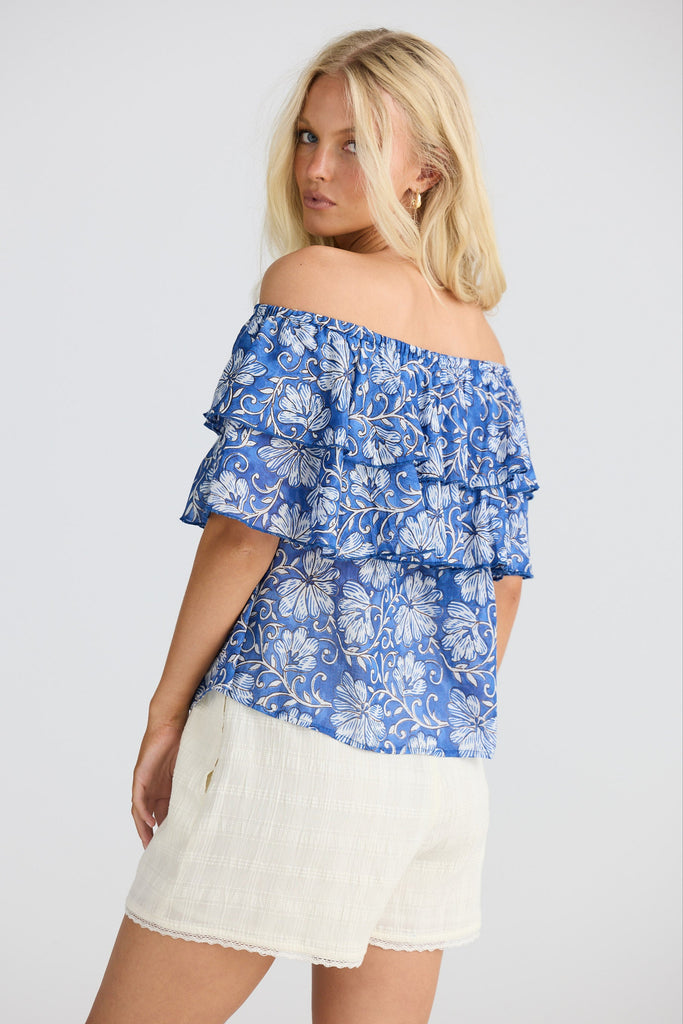 Woman wearing a blue floral off-shoulder top and white shorts on a plain background