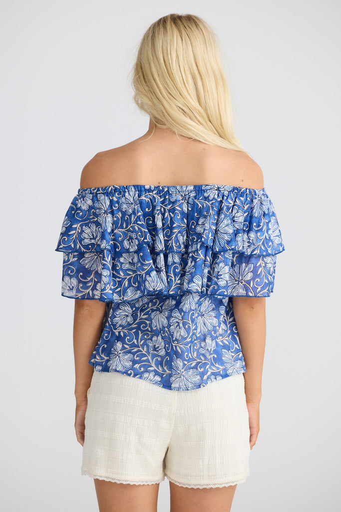 Woman wearing a blue floral off-shoulder top and white shorts on a plain background