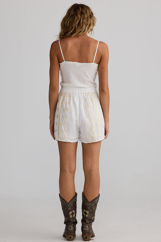 Woman wearing a white tank top and shorts with cowboy boots on a plain background