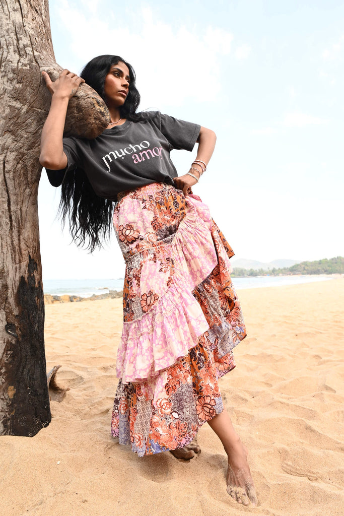 Person in a colorful dress and gray shirt standing on a beach with a tree in the background