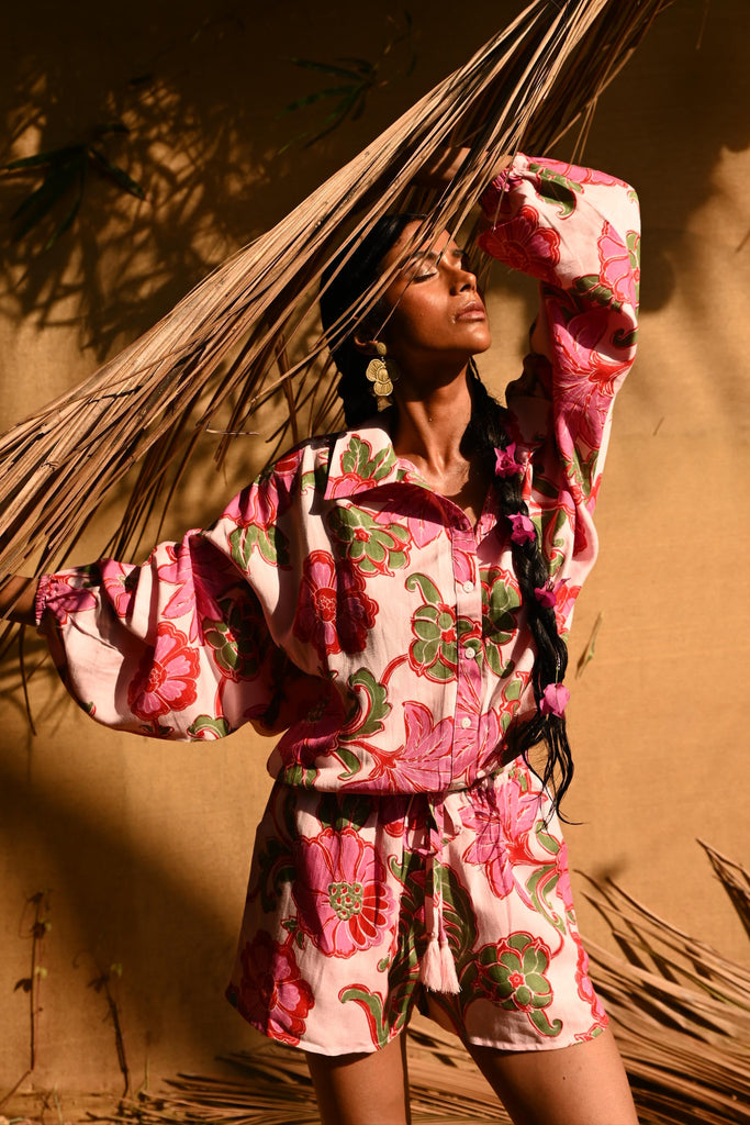 Person wearing pink floral shirt and shorts standing in front of a tan coloured wall with dried palm leaves.