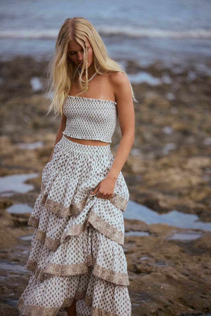 Woman in a patterned strapless top and tiered skirt standing on a beach with ocean in the background