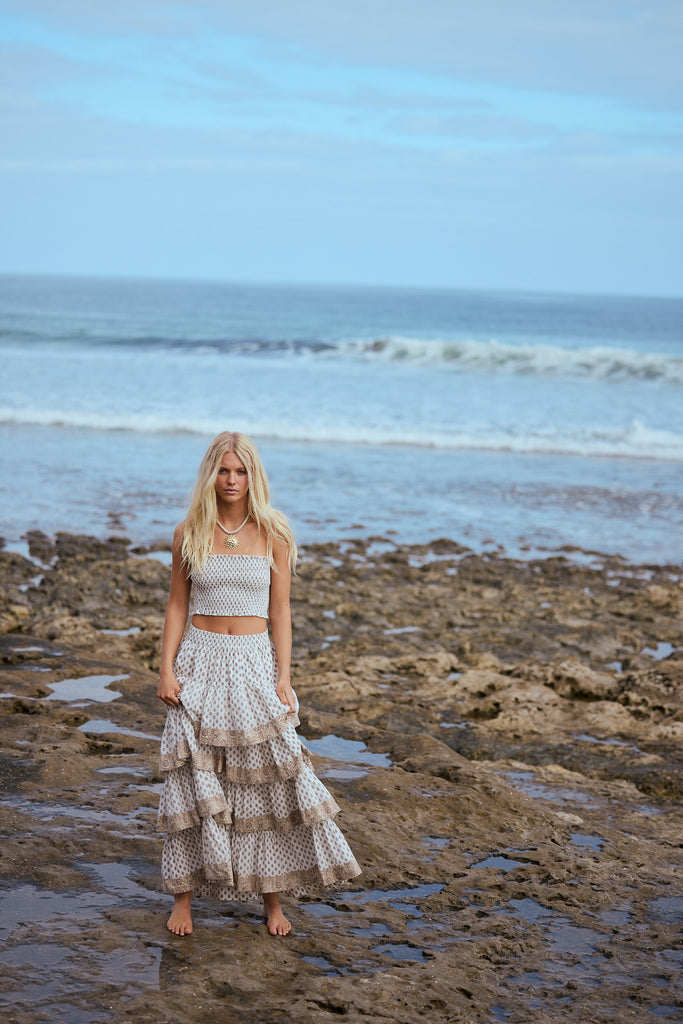 Woman in a patterned strapless top and tiered skirt standing on a beach with ocean in the background