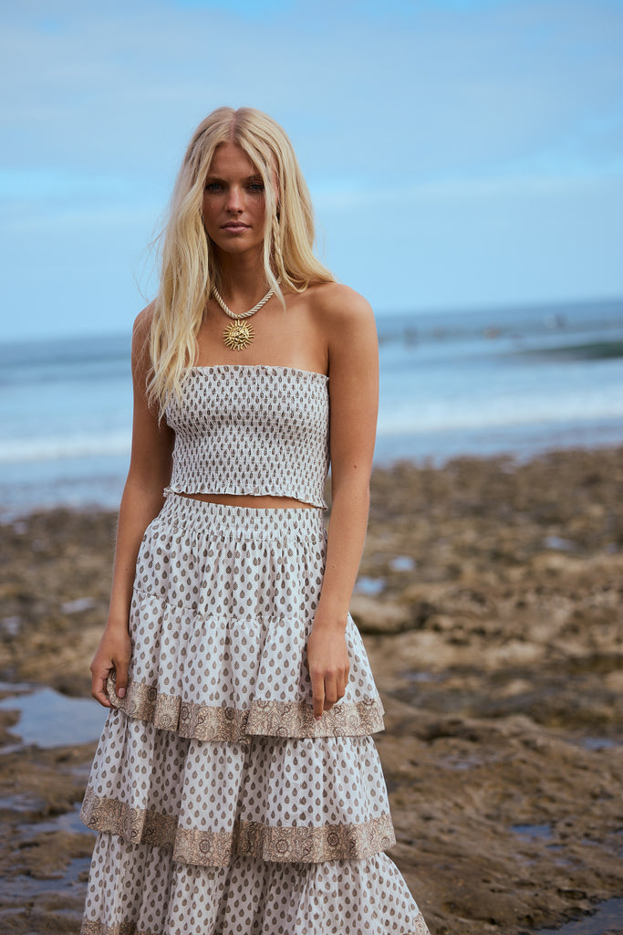 Woman in a patterned strapless top and tiered skirt standing on a beach with ocean in the background