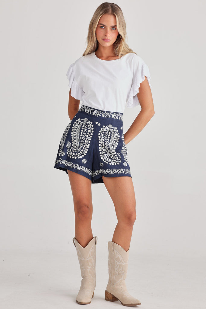 Woman wearing a white top and navy shorts with a pattern, standing against a plain background