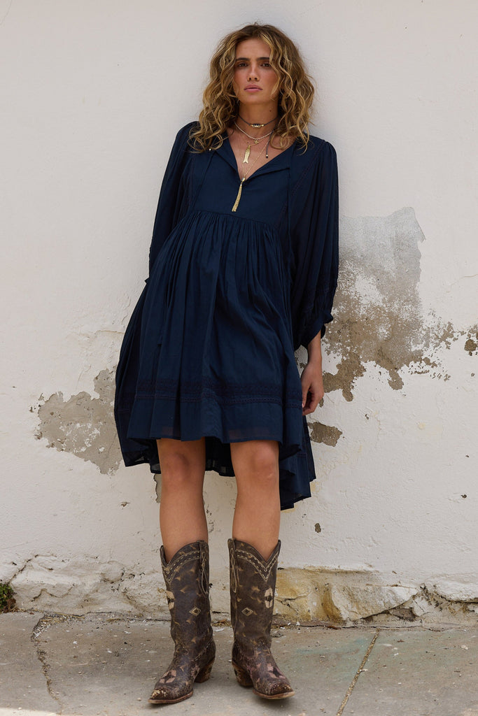 Woman wearing a navy dress and brown boots standing against a white wall.