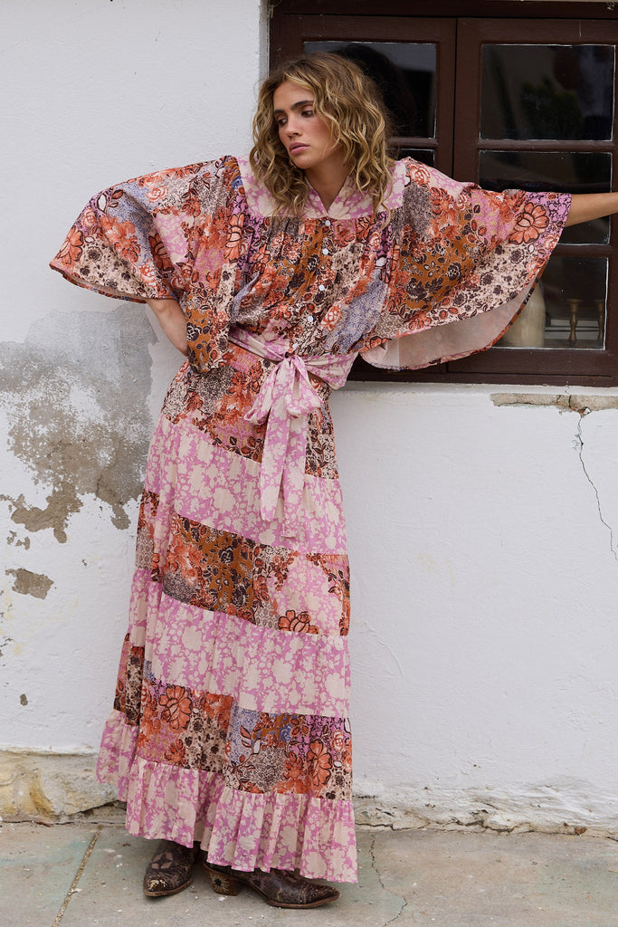 Woman wearing a floral top and skirt standing against a white wall with a window.