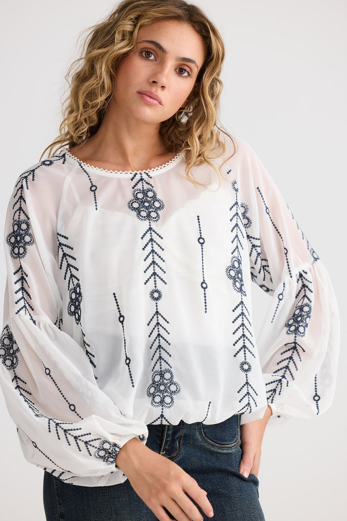 Woman wearing a white blouse with black floral and arrow patterns on a plain background