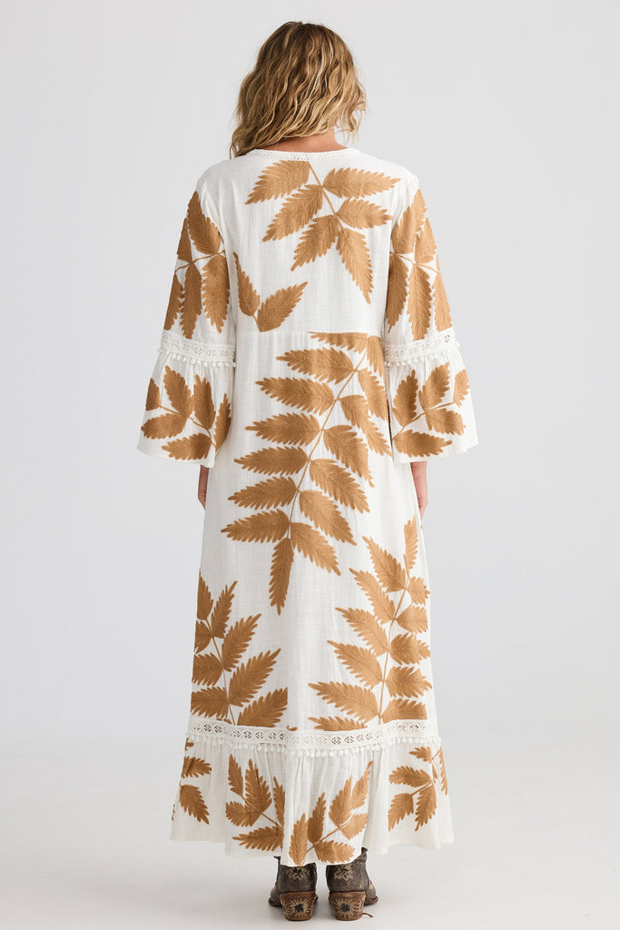 Woman wearing a long, white dress with brown leaf patterns on a plain background