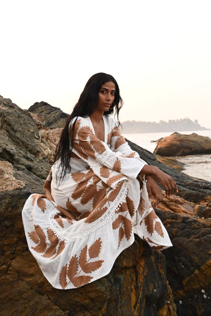 Woman in a white dress with brown patterns standing against a rocky cliff.