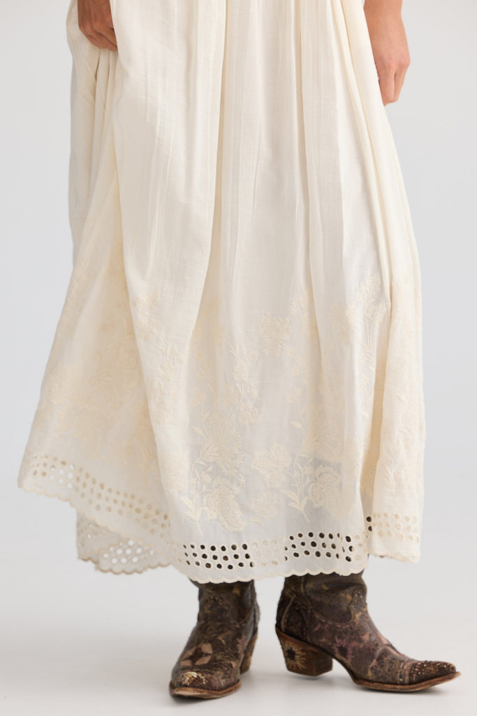 Person wearing a long off white dress with lace details and brown boots on a plain background