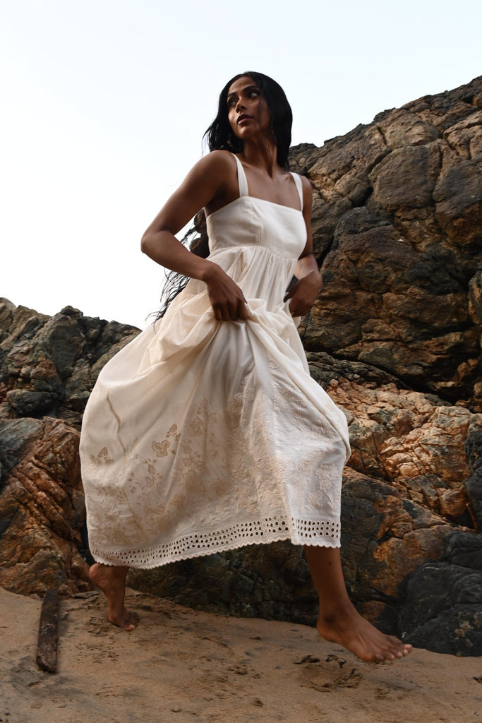 Woman in an off white dress running on a beach with rocky terrain and a clear sky.