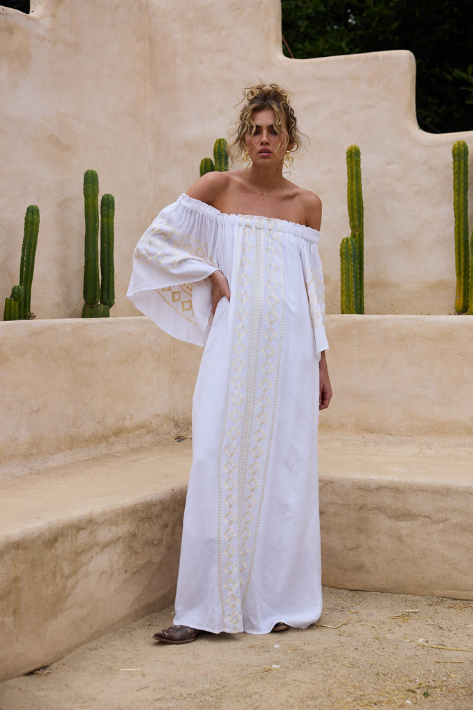 Woman in a white off-shoulder dress standing in front of a beige wall with cacti.