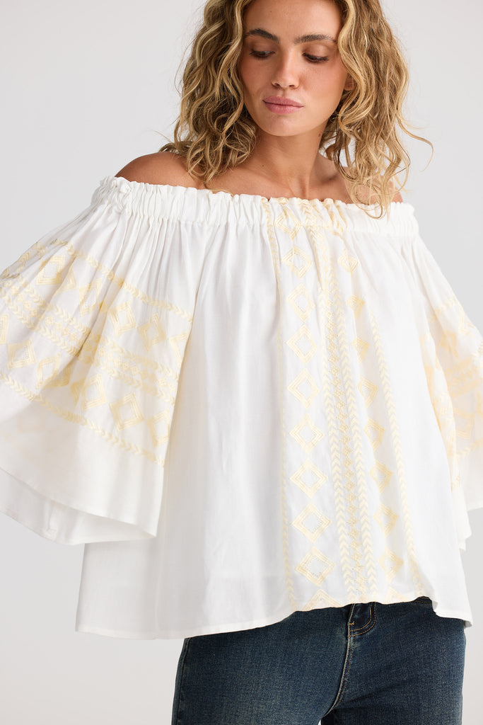 Woman wearing a white off-shoulder top with cream patterns and blue jeans on a plain background